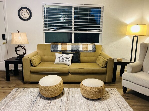 A living room with a mustard-colored sofa, two round ottomans, a 'welcome home' pillow, a clock, and a lamp on a side table. - Giddy Up Getaway in Nampa (Nampa)