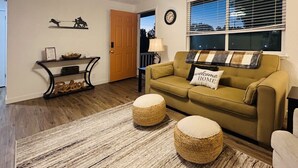Un salon chaleureux avec un canapé, deux poufs, une table basse, une étagère décorative et un ventilateur de plafond.