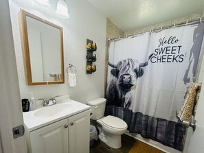 A bathroom with a white sink, mirror, and toilet. There is a shower curtain with a cow design and the text "Hello SWEET CHEEKS". - Giddy Up Getaway in Nampa (Nampa)