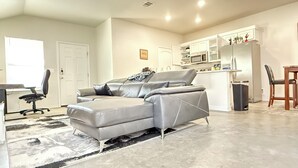 A modern living room with a grey sectional sofa, a dining area with a wooden table and chairs, and a kitchen with white cabinets and appliances.
