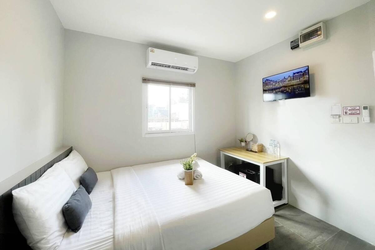 A bedroom with a bed, a small bedside table with a plant, a window, an air conditioner, a TV, and a wall-mounted heater.