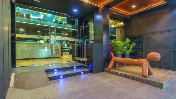 A modern hotel entrance with a glass facade, a metal bench, and a potted plant. - Deco Lux Hotel (Bangkok)