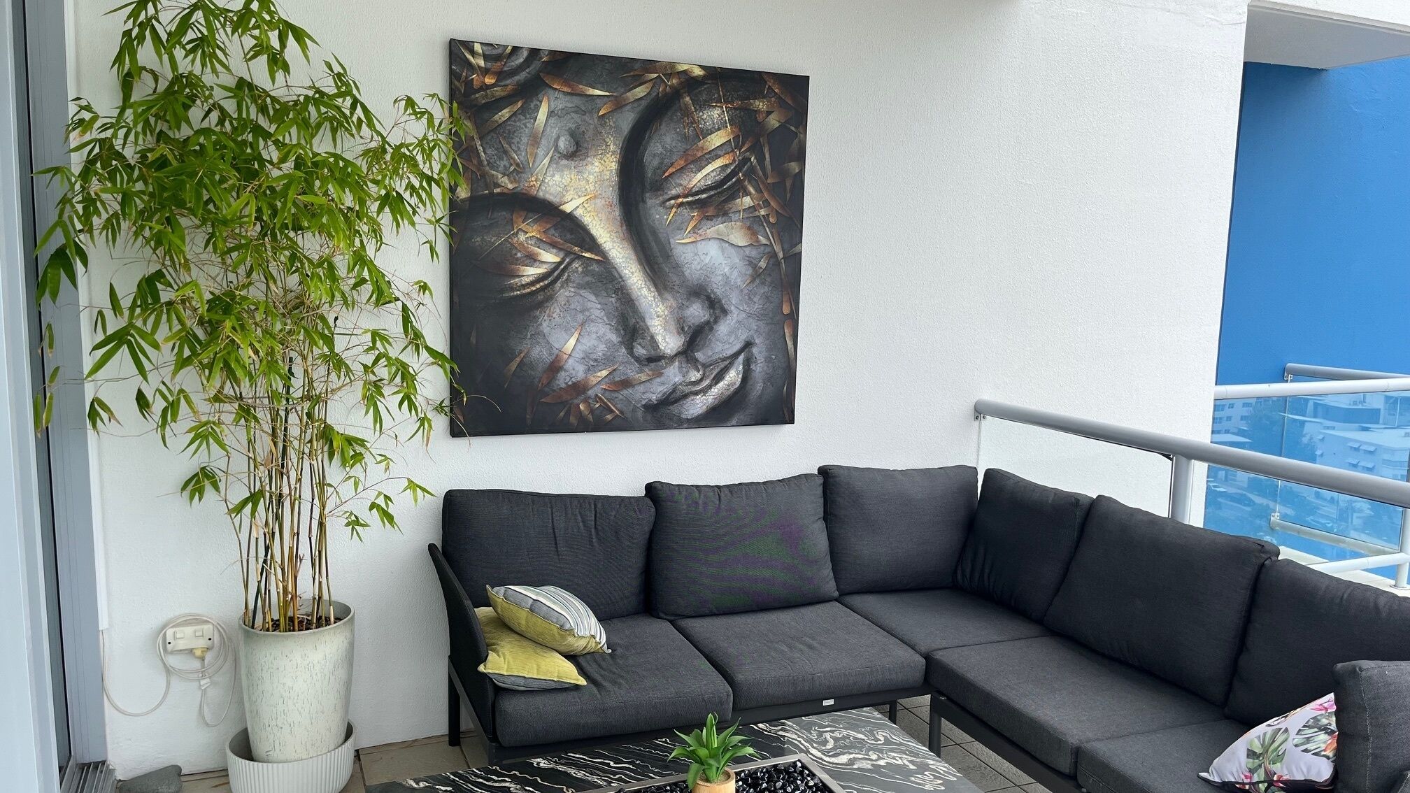 Een balkon met een zwarte modulaire bank, een groot abstract schilderij, een pot met bamboe en uitzicht op een blauw gebouw.