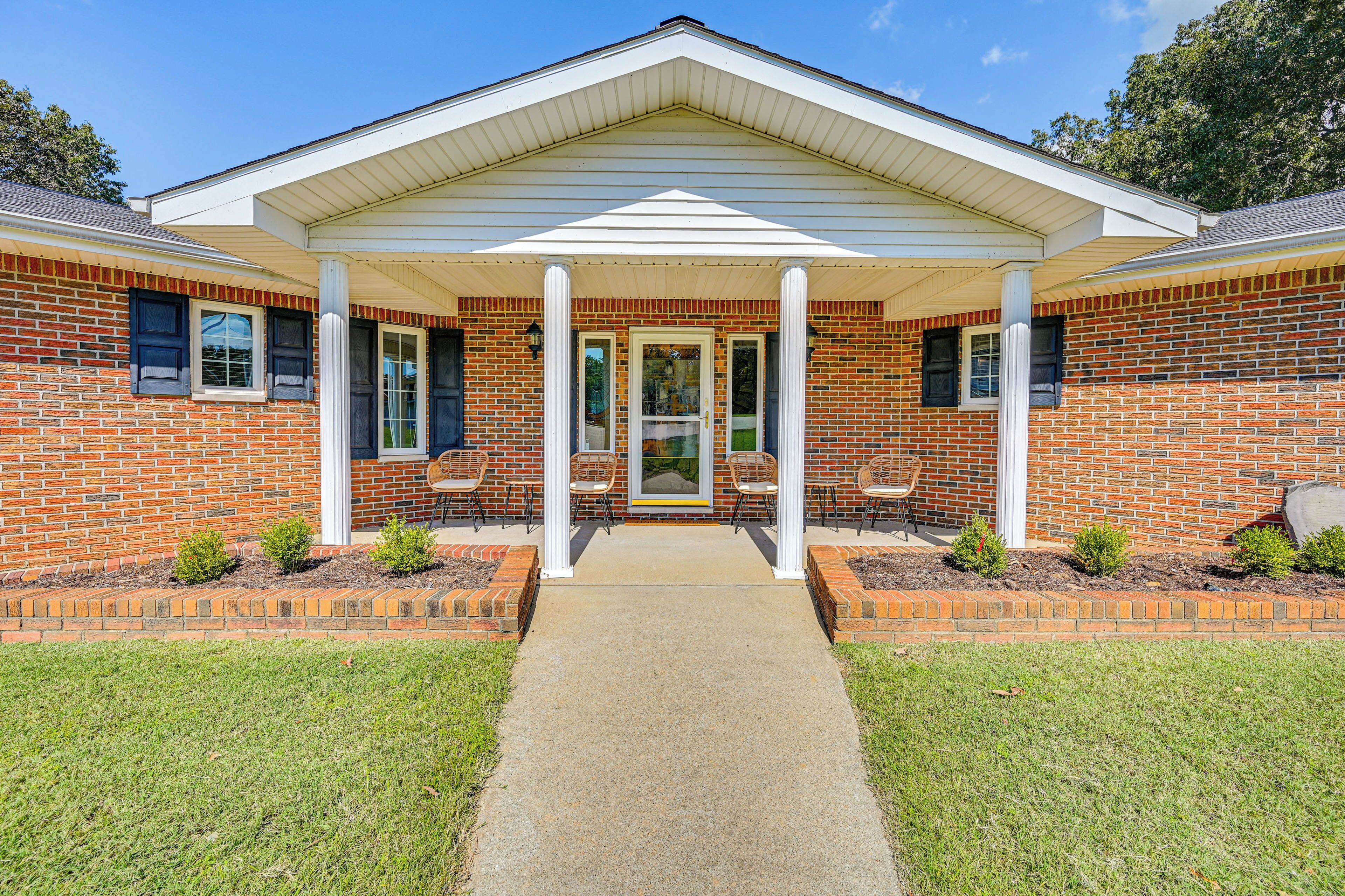A brick building with a covered porch, a concrete pathway, and small shrubs.
