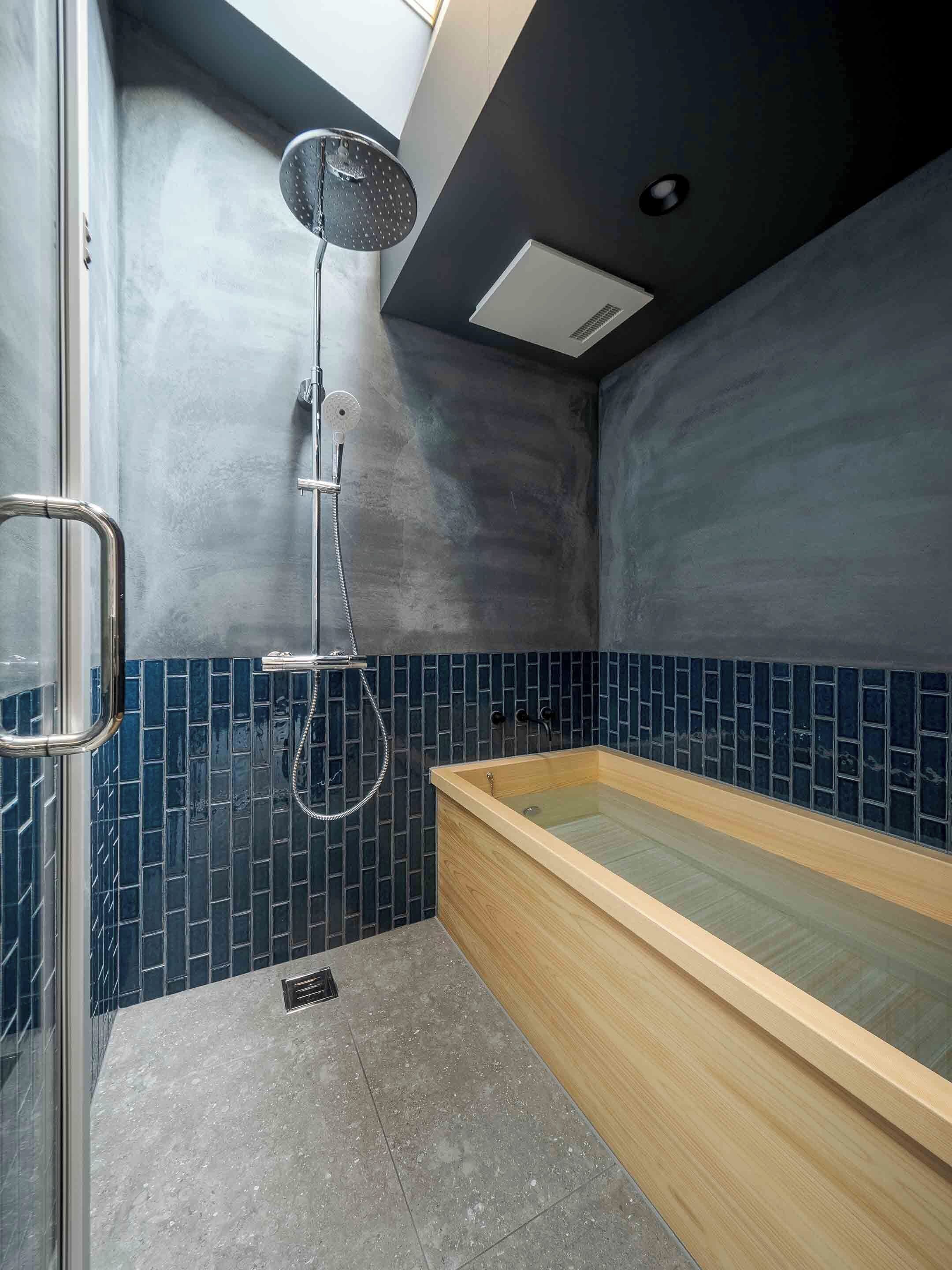 A modern bathroom with a wooden bathtub, a glass shower enclosure, and dark blue mosaic tiles.