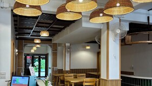 A restaurant interior with wicker pendant lighting, wooden tables and chairs, a reception desk with a computer, and a patterned floor. - Charm Hostel at Jomtien (Pattaya)