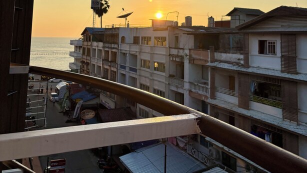 A balcony with a view of a sunset over the sea, buildings in the background, and a satellite dish on one of the buildings. - Charm Hostel at Jomtien (Pattaya)