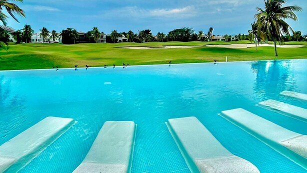 Une piscine d’un bleu éclatant, dotée de chaises longues, entourée d’une pelouse bien entretenue et de palmiers, sous un ciel partiellement nuageux.