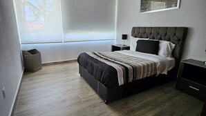 Une chambre à coucher moderne avec un grand lit, une table de chevet, une fenêtre avec des stores et un tableau encadré au mur.