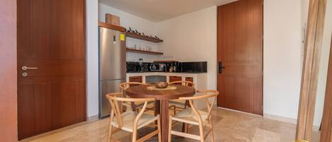 A dining area with a round wooden table and four chairs, a kitchen with a refrigerator and shelves above the counter, and wooden doors.