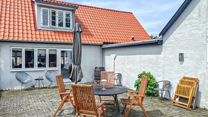 A patio with a round table and chairs, a covered grill, and a white wall with a window. - Nice home in Rønne with WiFi (Rønne)