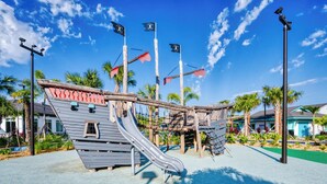 Un terrain de jeux avec un toboggan et des structures d’escalade, entouré de palmiers et surplombé d’un ciel bleu azur.
