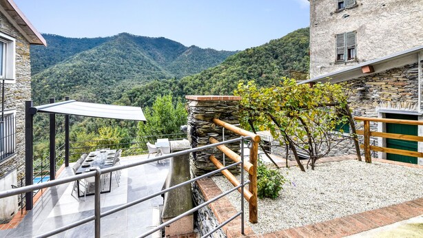Una terraza con una mesa y sillas para comer, un muro de piedra y vistas a una montaña arbolada.
