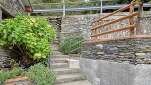 Una escalera de piedra con barandilla de metal, rodeada de follaje y un muro de piedra.