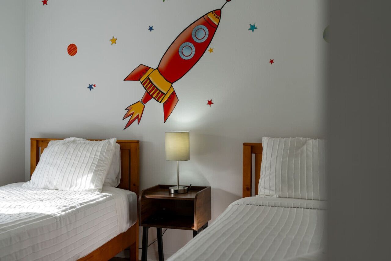A hotel room with two beds, a nightstand with a lamp, and a wall mural of a rocket.