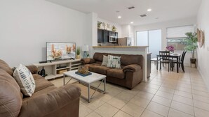 A modern living room with a brown sofa, a coffee table, and a TV. There is a kitchen area with cabinets and a dining table with chairs. - Cozy 3BDR House near Disney - AL (Kissimmee)