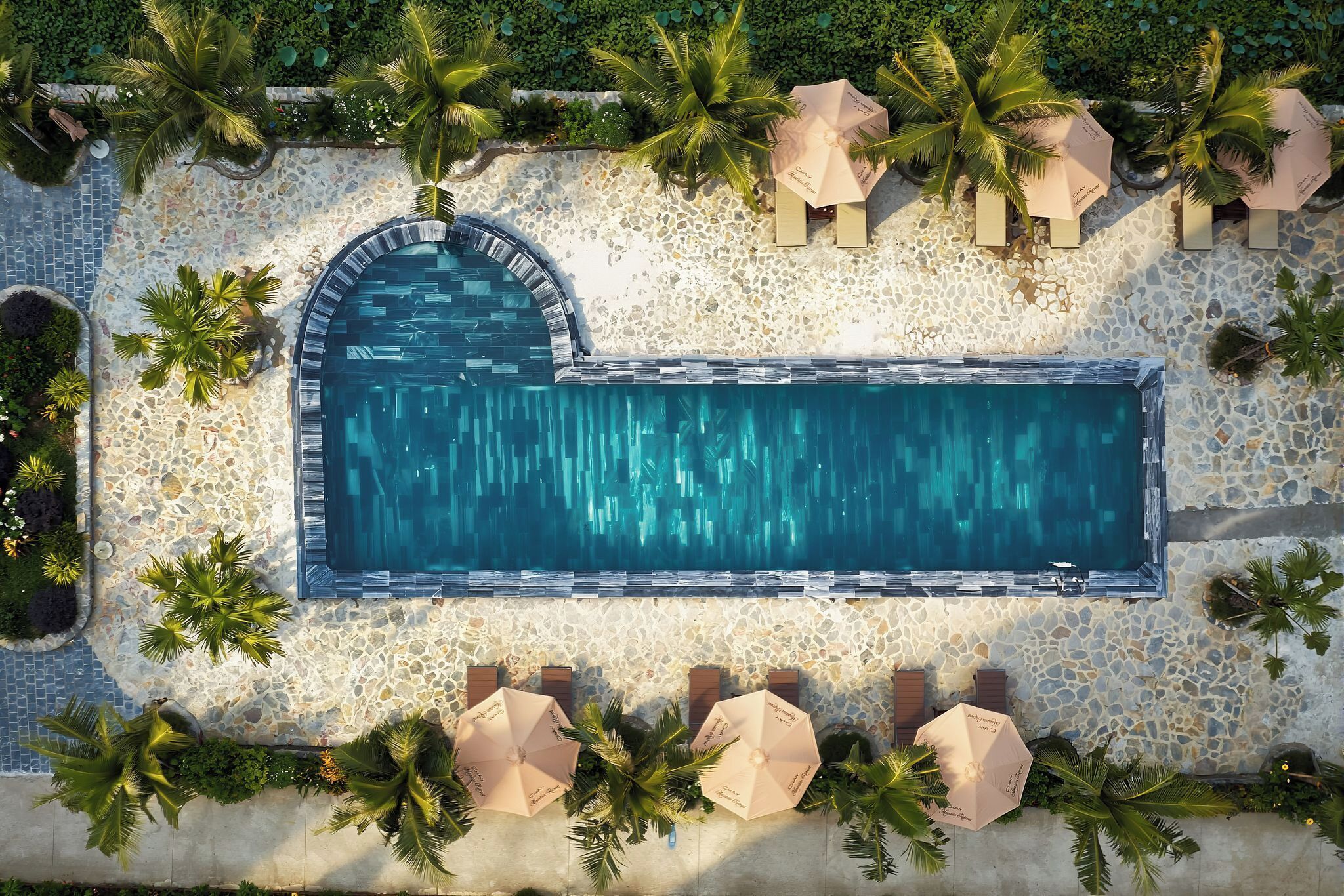 Ein Luftbild von einem rechteckigen Schwimmbecken, umgeben von einer Terrasse mit Kieselmuster, Palmen und Sonnenschirmen.