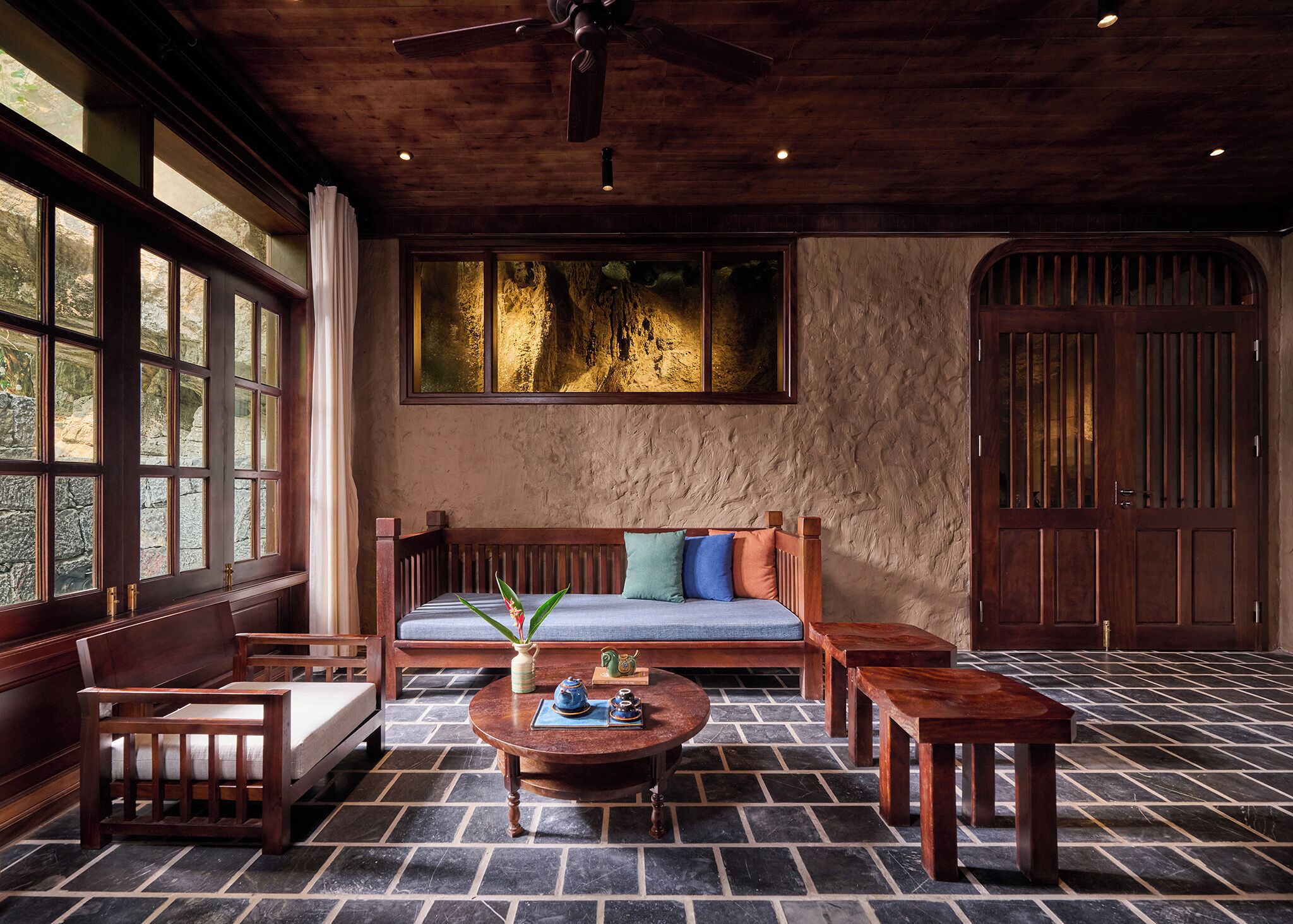 A room with a stone-tiled floor, a wooden sofa with cushions, a round wooden coffee table, and a large window with a view of trees.