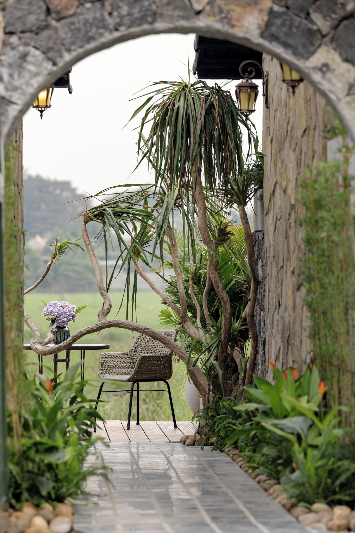 Un área de estar al aire libre con un arco de piedra, una silla de mimbre, una mesa con un jarrón de flores y vistas a un paisaje verde.