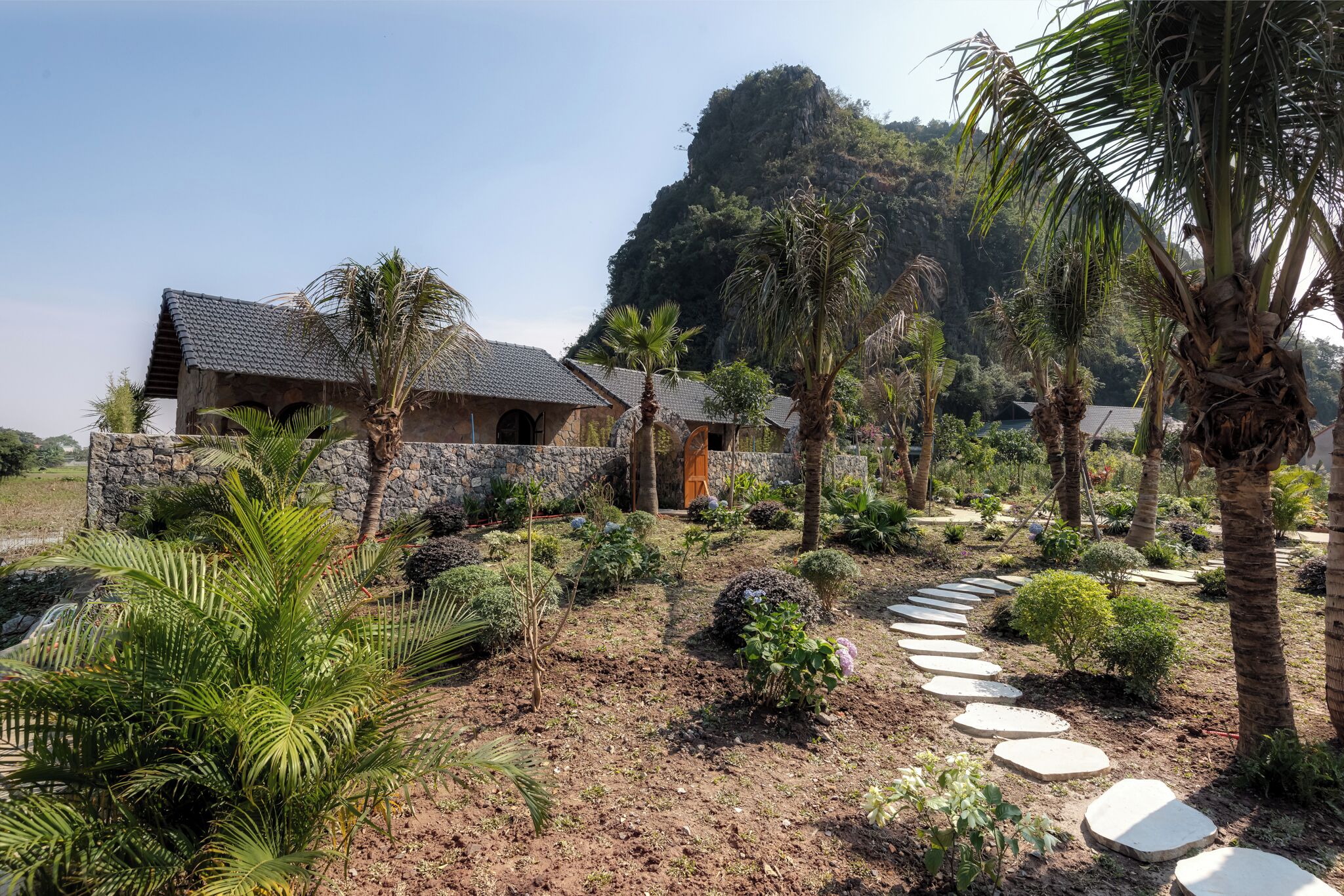 A rustic building with stone walls and a thatched roof, surrounded by a garden with palm trees and a variety of plants.