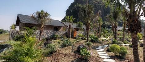 A rustic building with stone walls and a thatched roof, surrounded by a garden with palm trees and a variety of plants.