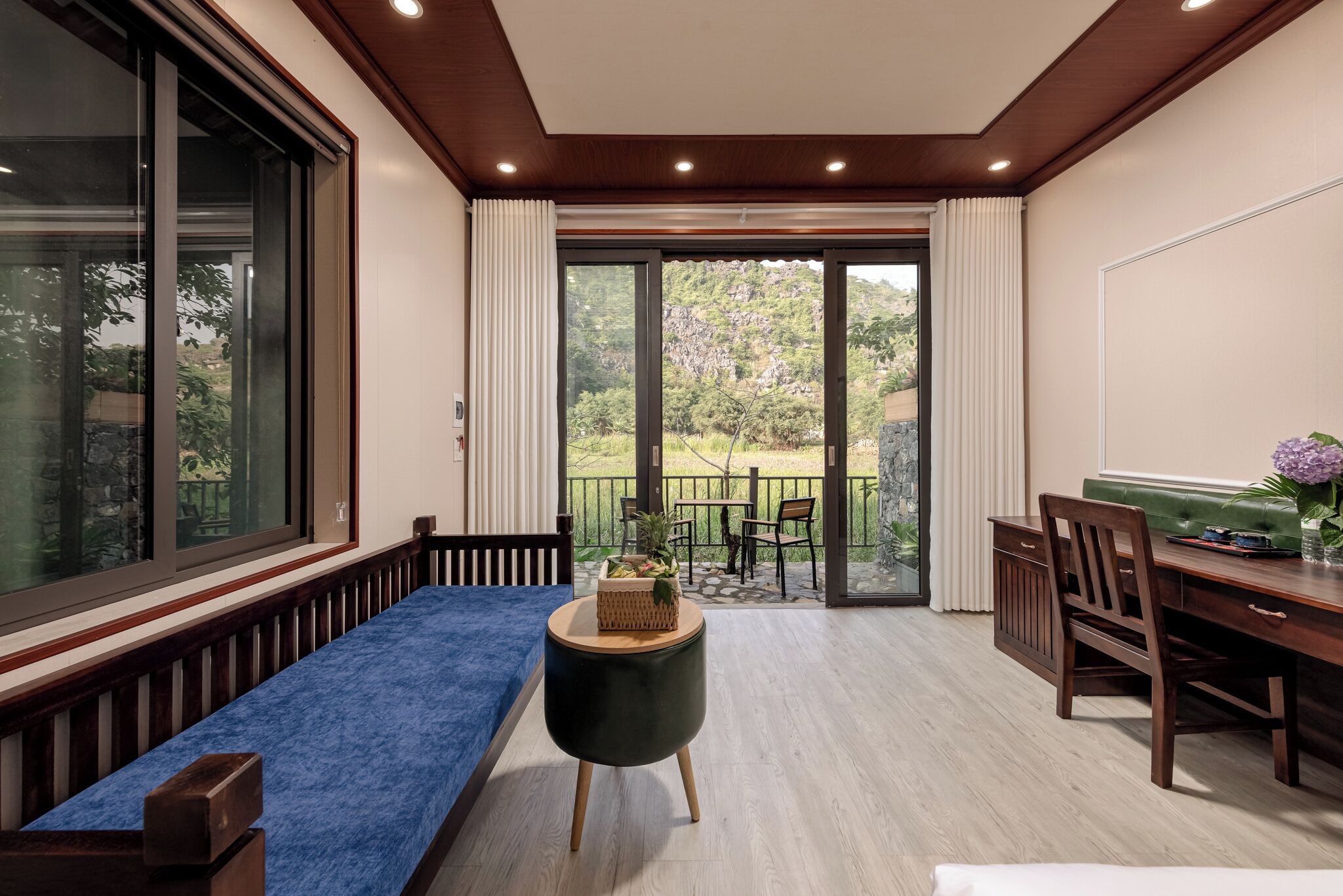 Una habitación con un sofá azul, una mesa de comedor de madera y un taburete pequeño y redondo. Hay una puerta corrediza de vidrio que da a un espacio exterior con vistas a zonas verdes y una terraza.