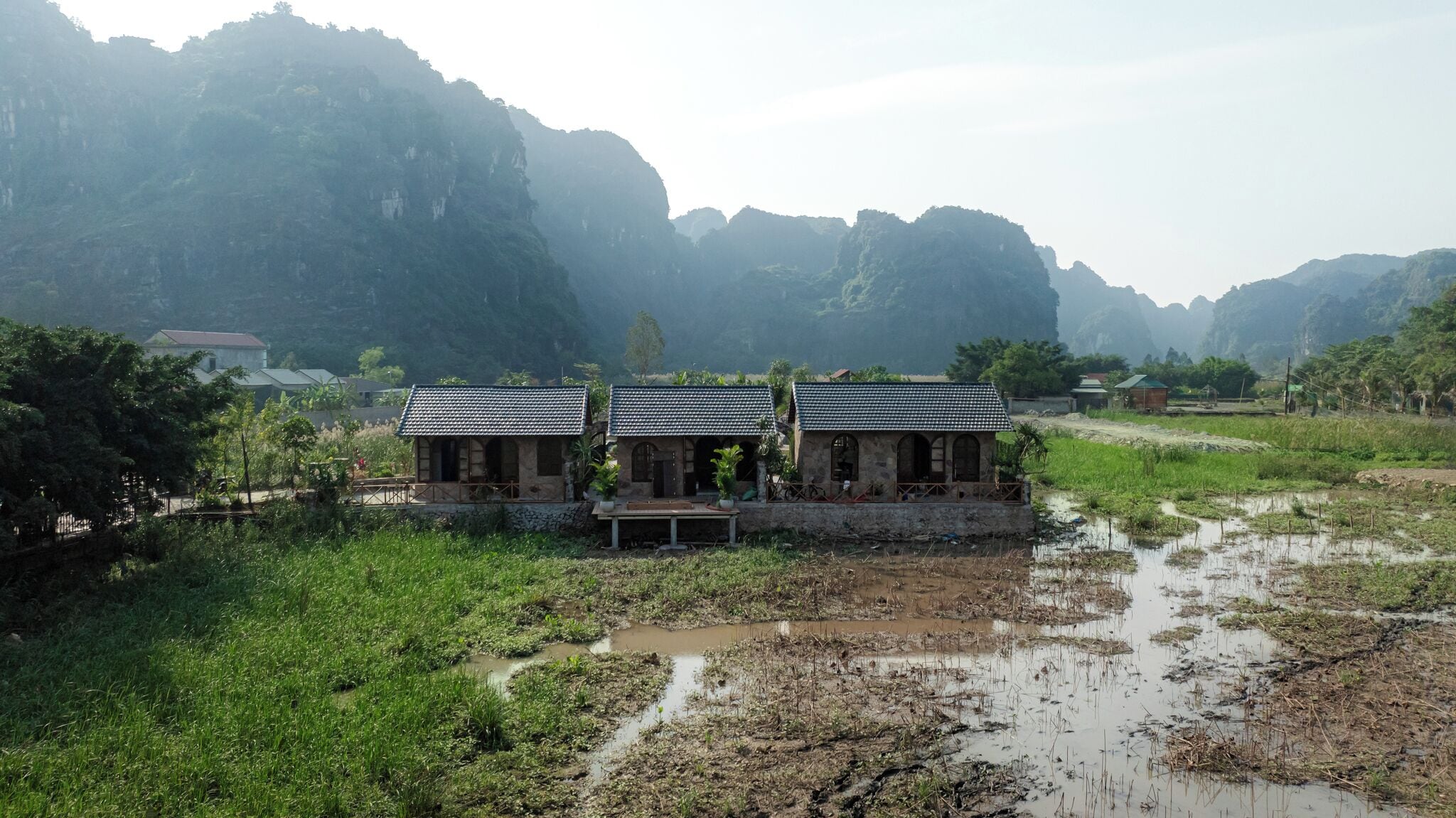 Ein Landhaus mit Ziegeldach, umgeben von üppiger Vegetation und einem Reisfeld.