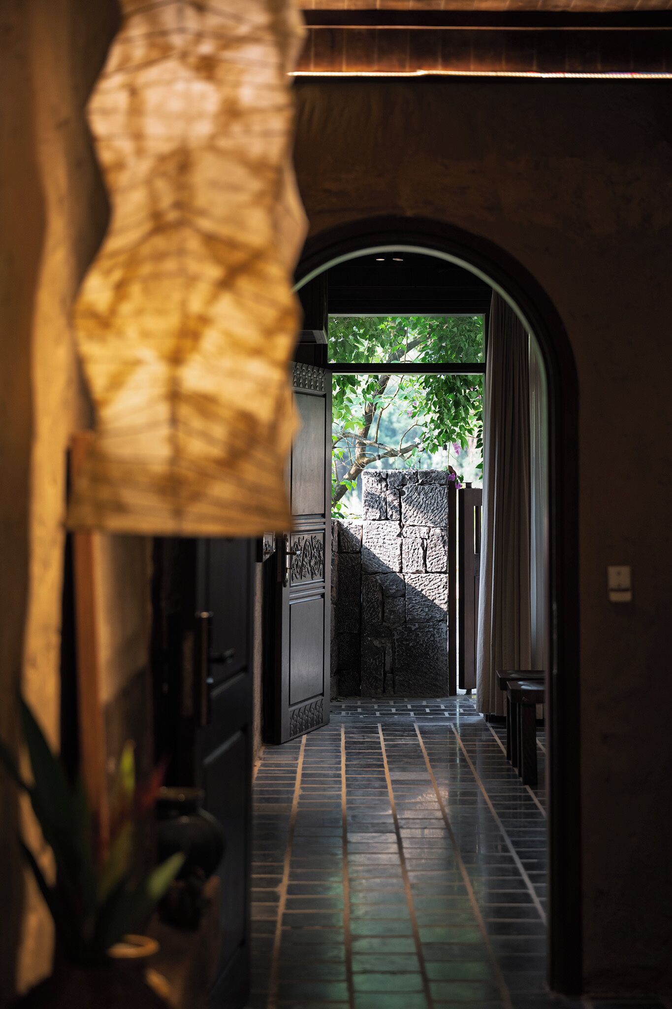 A hallway with a tiled floor, an arched doorway leading to a garden, and a hanging light fixture.