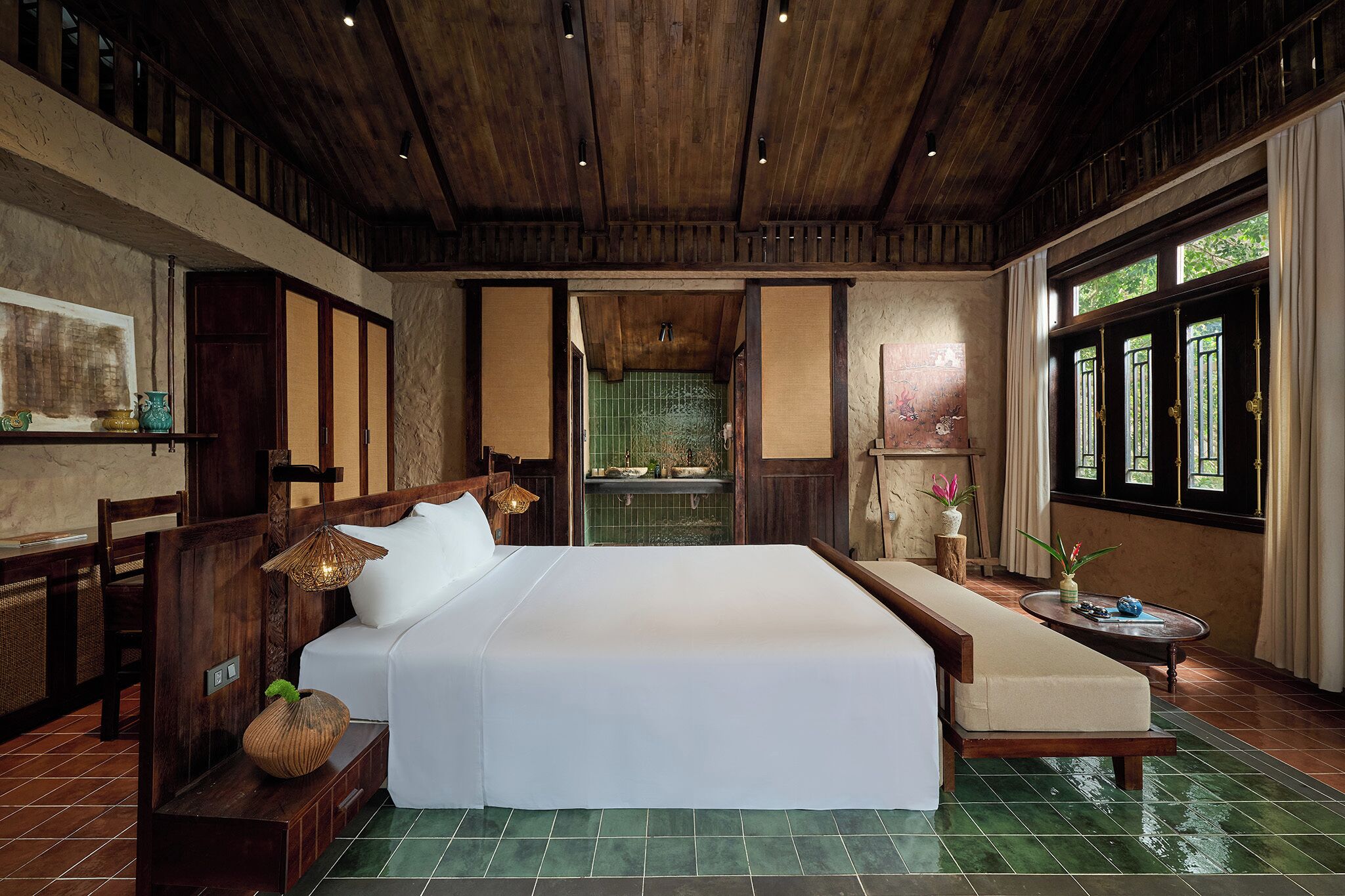 Una habitación con una cama, un sofá, una pequeña mesa con objetos decorativos, una ventana con cortinas y vistas a una zona con azulejos verdes.