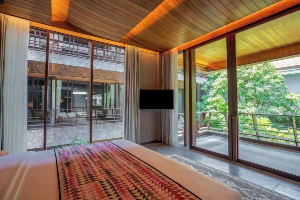 A modern hotel room with a large bed, a flat-screen TV, and a balcony with a view of greenery.