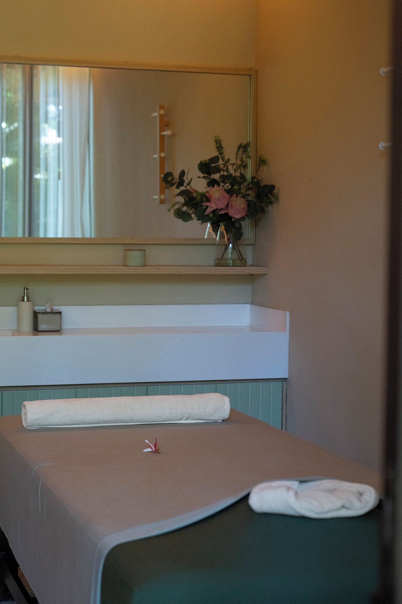 A room with a bed, a small table with a towel, a mirror, and a vase with flowers.