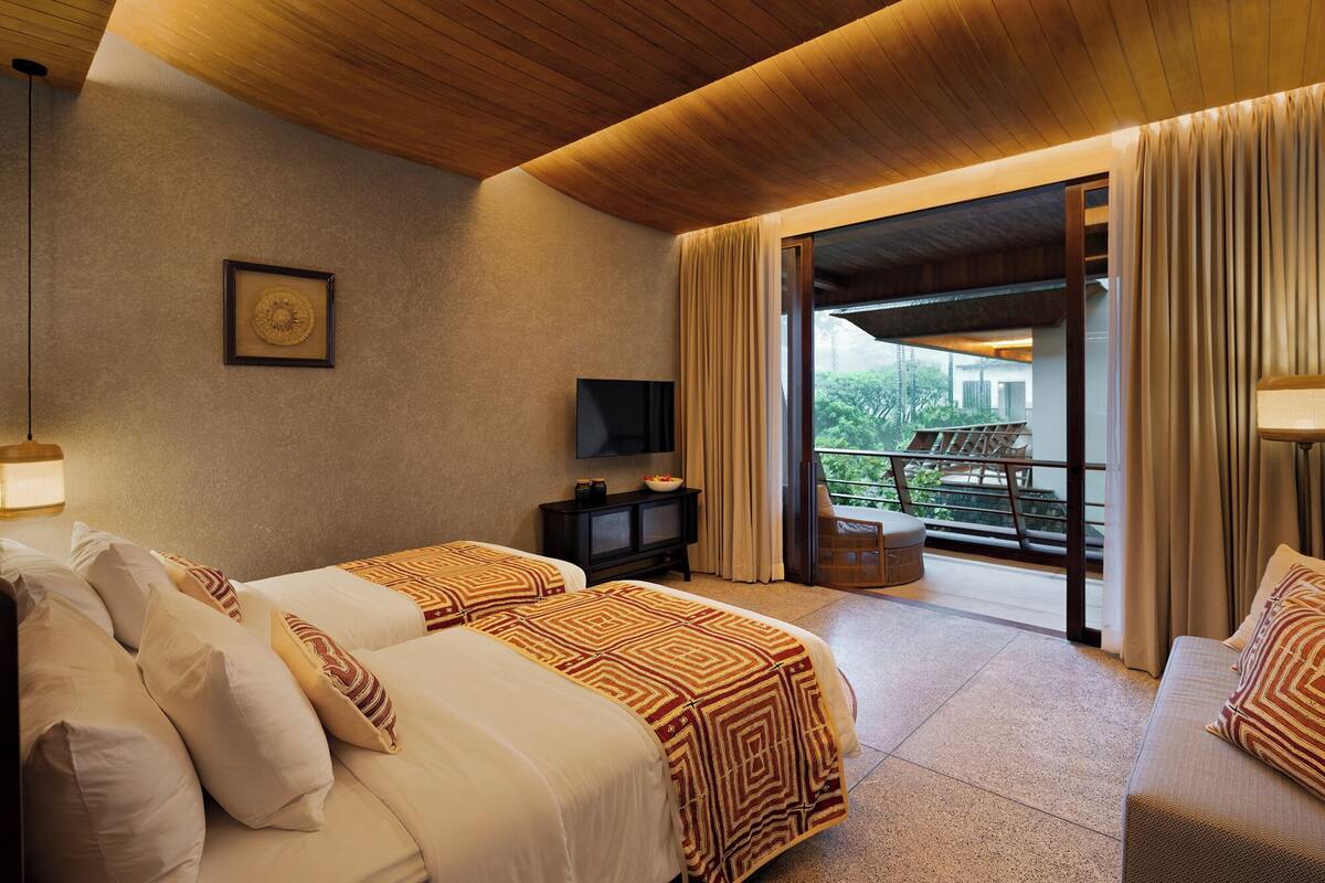 A hotel room with two beds, a TV, a small table with a bowl of fruit, and a balcony with a view of greenery.