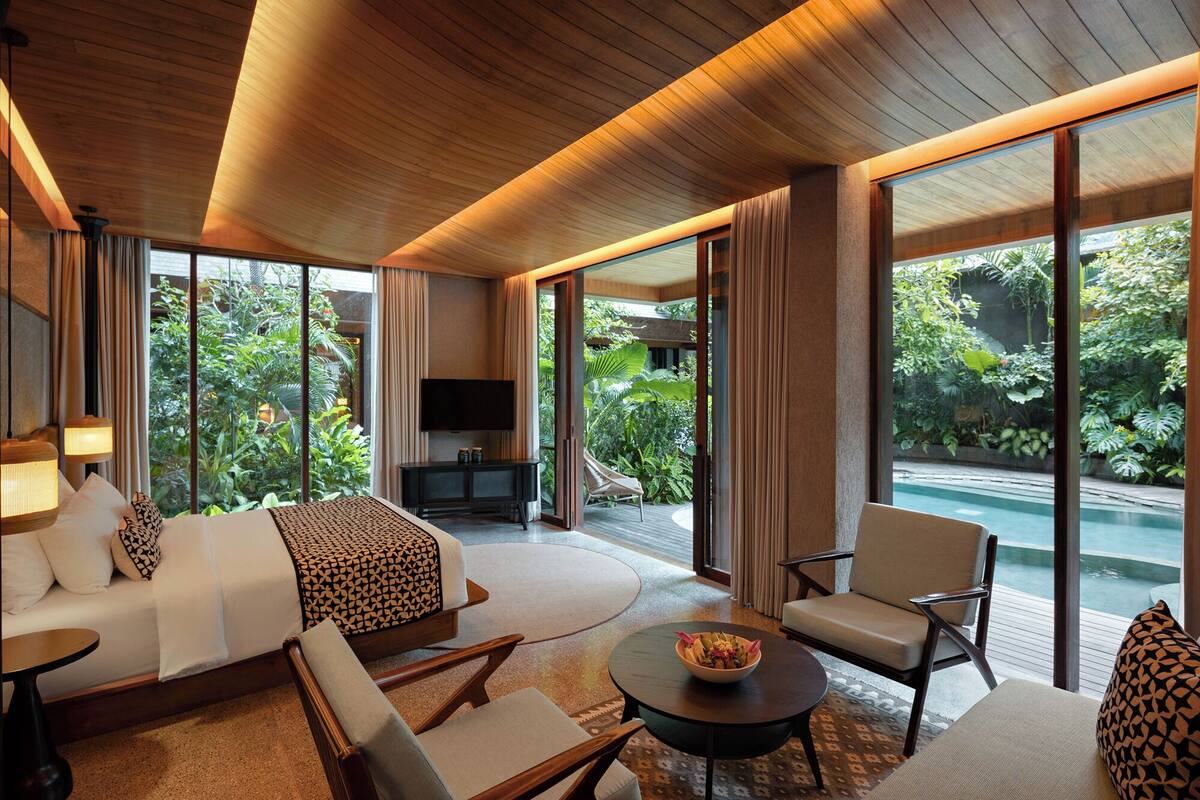 A modern hotel room with a large bed, seating area, and a view of a pool and garden.