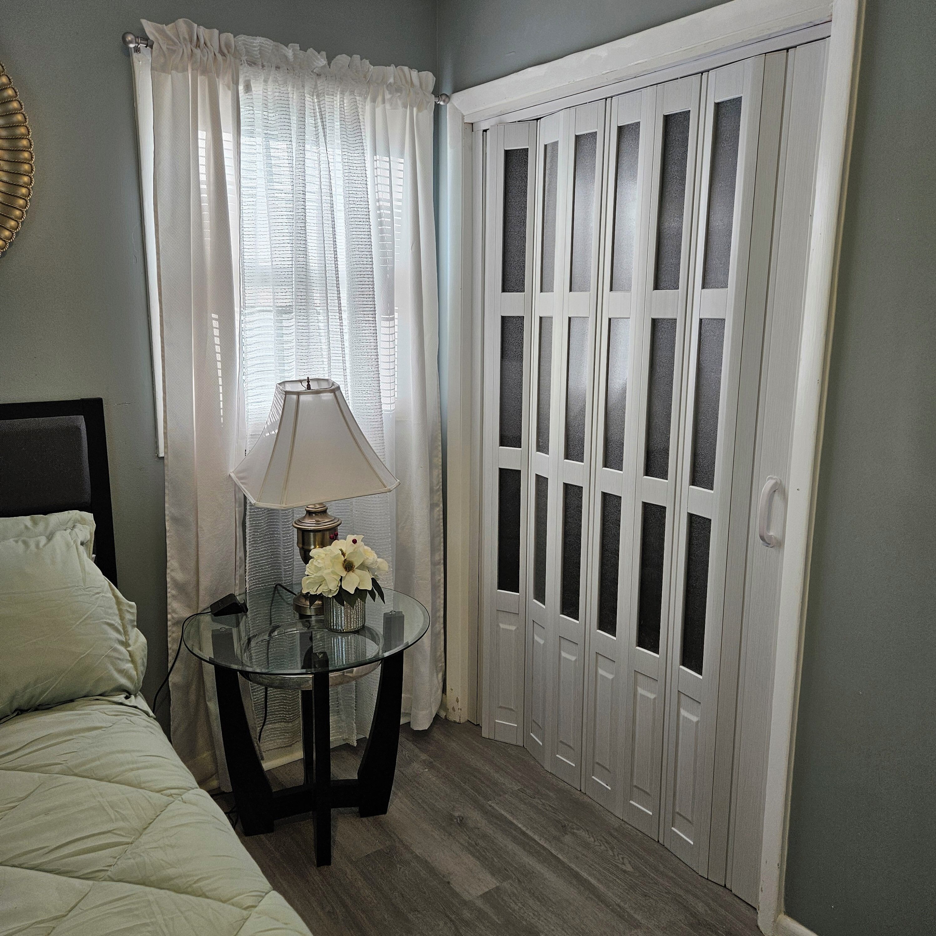 A bedroom with a bed, a glass table with a lamp and flowers, a window with curtains, and a set of white doors.