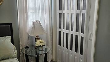 A bedroom with a bed, a glass table with a lamp and flowers, a window with curtains, and a set of white doors.