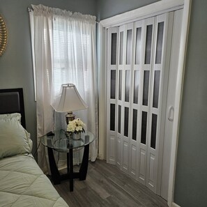 A bedroom with a bed, a glass table with a lamp and flowers, a window with curtains, and a set of white doors.