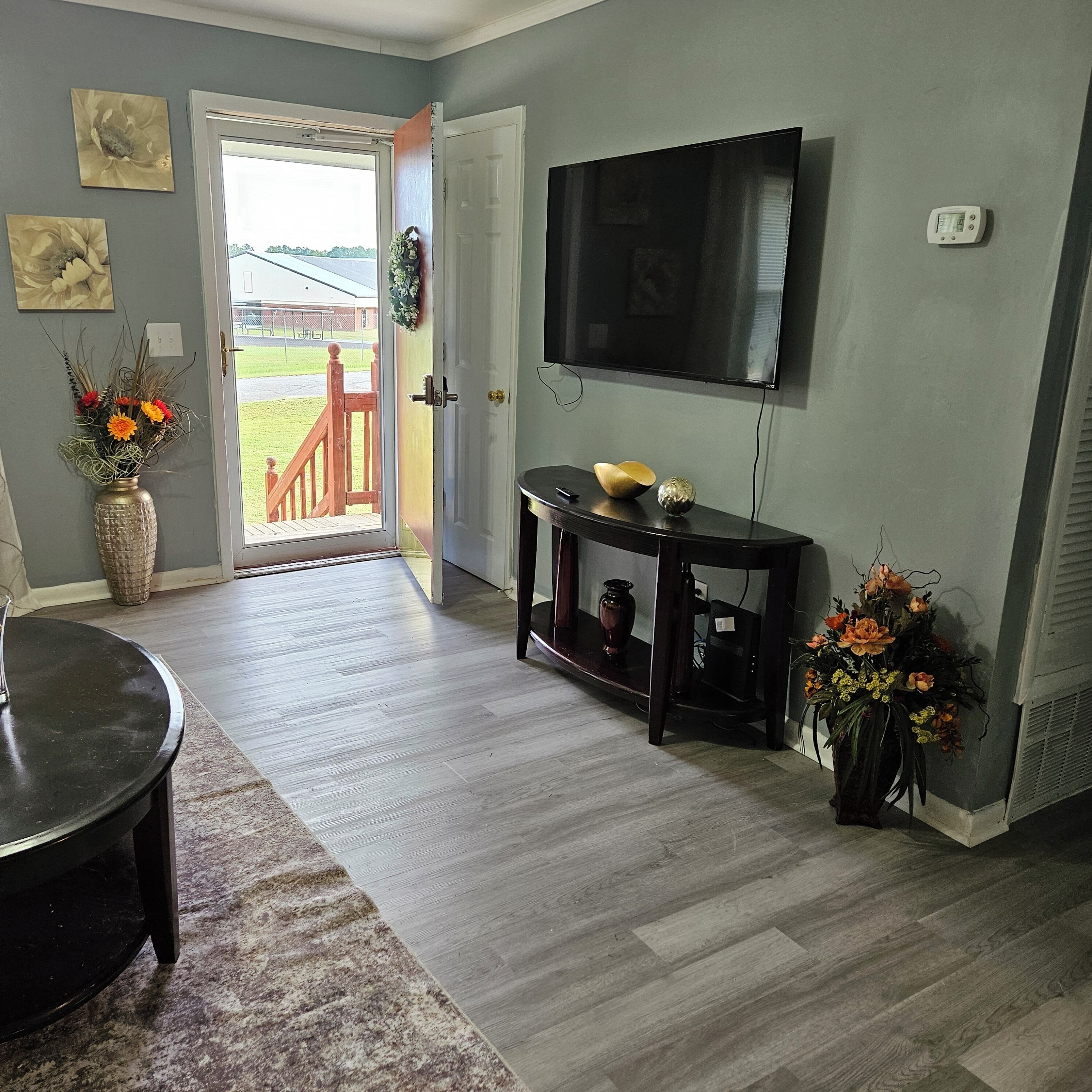 A living room with a TV mounted on the wall, a wooden coffee table, a rug, and a door leading outside.