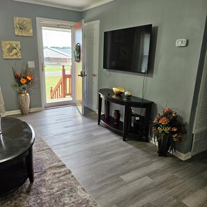 A living room with a TV mounted on the wall, a wooden coffee table, a rug, and a door leading outside.