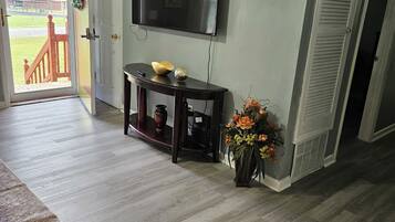 A living room with a flat-screen TV mounted on the wall, a console table with decorative items, a potted plant, and a view of the outdoors through an open door.