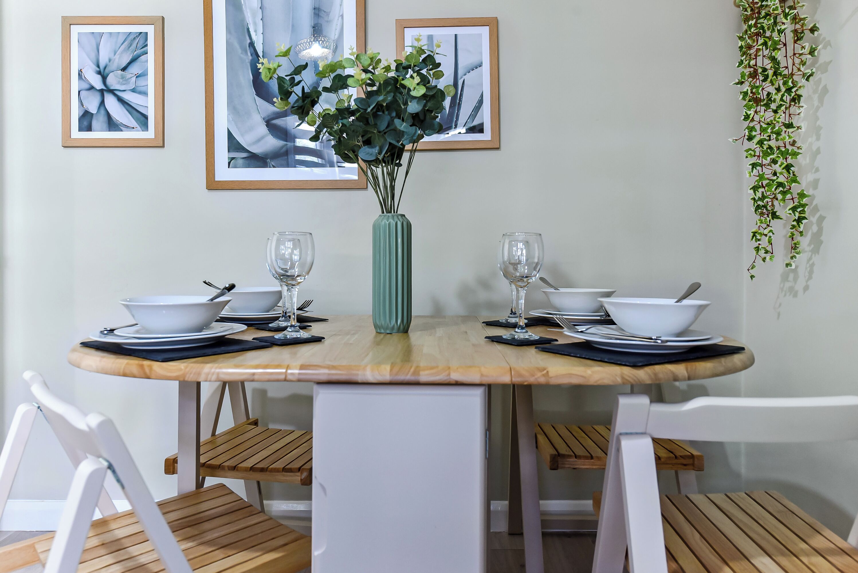 A dining table set with white bowls, wine glasses, and cutlery, flanked by framed pictures and a hanging plant.