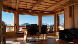 A wooden interior with a dining table and chairs, large windows with mountain views, and a black sofa.