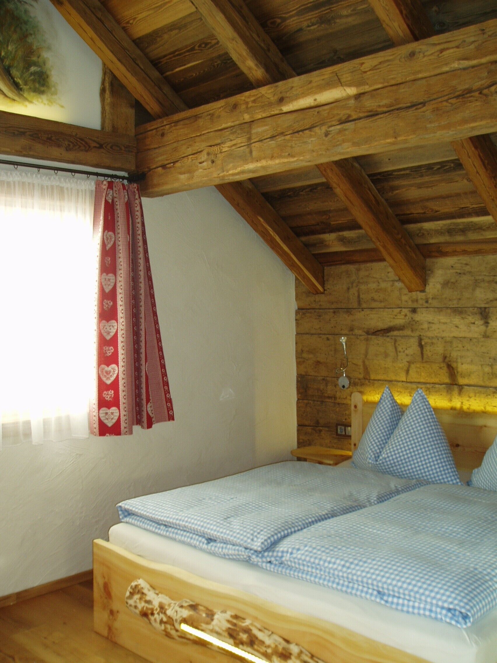 Una camera da letto con soffitto in legno, un letto con copriletto a quadretti, una finestra con tende a cuoricini e una testiera in legno.
