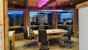 A wooden interior with a large table and chairs, a kitchen visible through the windows, and a ceiling with exposed beams.