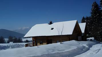 A log cabin with a snow-covered roof, surrounded by snow-covered ground and trees, with a clear sky in the background.