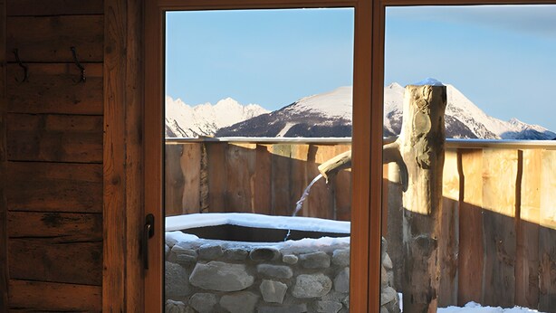 A wooden cabin interior with a stone hot tub outside, a wooden door, and a view of snow-covered mountains.