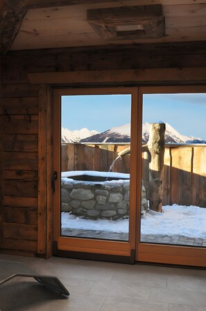 A wooden cabin interior with a stone hot tub outside, a wooden door, and a view of snow-covered mountains.
