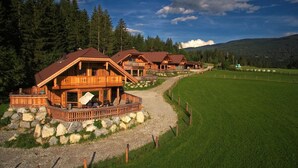 A row of wooden cabins with stone foundations, set in a green field with a gravel path leading to them.