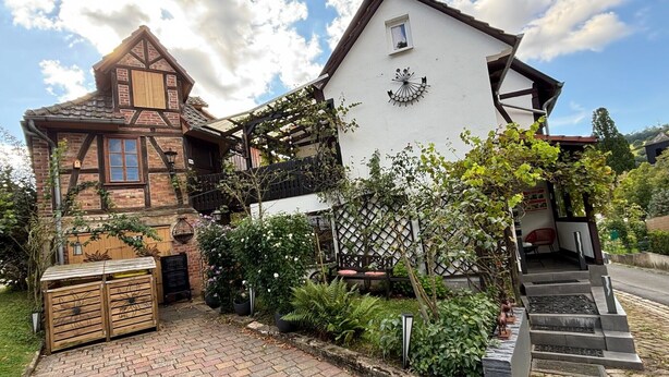 Una casa tradicional de entramado madera con sección superior blanca, sección inferior de ladrillo y un jardín con banco de madera y plantas en macetas.