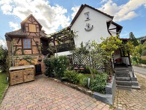 A traditional half-timbered house with a white upper section, a brick lower section, and a garden with a wooden bench and potted plants.