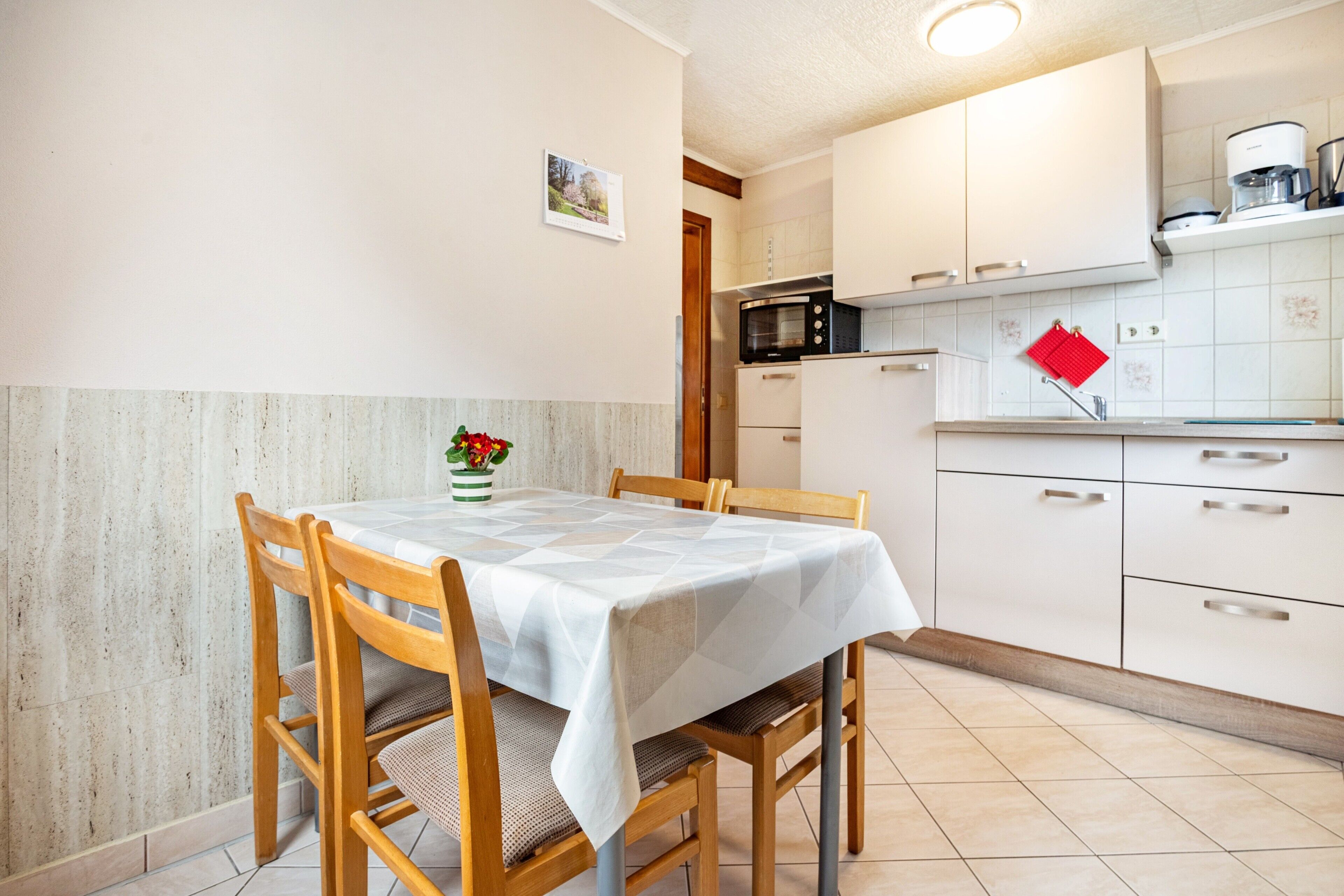 A dining area with a table set for four, wooden chairs, a white tablecloth, a vase with flowers, and a kitchen area with white cabinets and a red kettle.